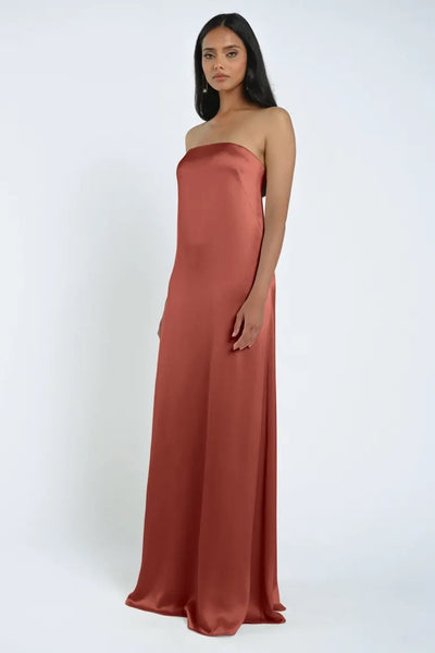 A woman stands against a plain background, wearing the strapless, floor-length James Bridesmaid Dress by Jenny Yoo in rust-colored luxe satin fabric from Bergamot Bridal.