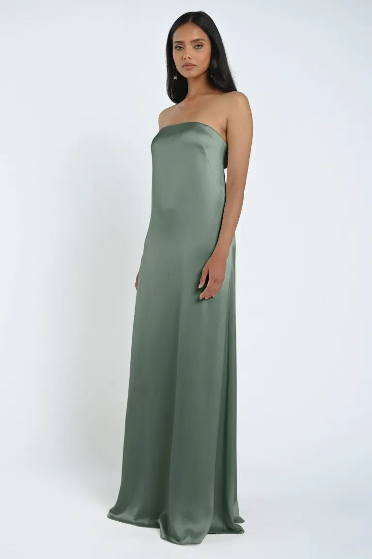 A woman stands wearing the James Bridesmaid Dress by Jenny Yoo from Bergamot Bridal, a strapless, floor-length, sage green gown made of luxe satin fabric in a sleek sheath silhouette against a white background.