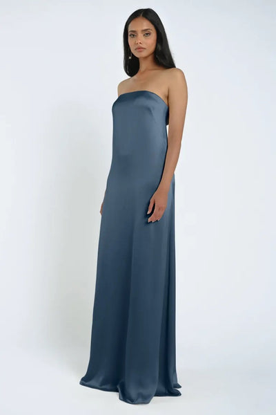 A woman stands wearing a long, strapless James Bridesmaid Dress by Jenny Yoo in blue luxe satin fabric with a graceful sheath silhouette against a plain light background, brought to you by Bergamot Bridal.