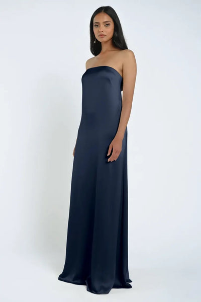 A woman stands wearing a strapless navy blue James Bridesmaid Dress by Jenny Yoo, crafted from luxe satin fabric, against a plain white background.