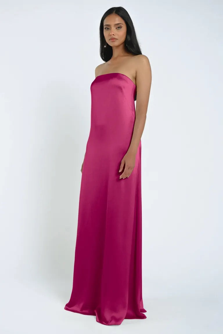 A woman stands facing the camera, wearing a strapless James Bridesmaid Dress by Jenny Yoo from Bergamot Bridal, made from luxe satin fabric in a stunning magenta hue, all set against a plain white background.