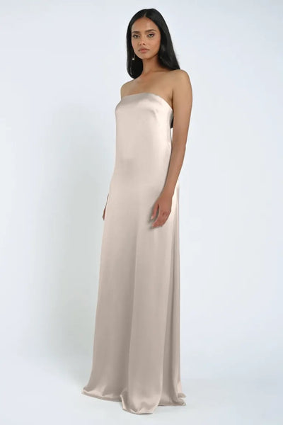 A woman stands wearing the James Bridesmaid Dress by Jenny Yoo, a strapless, floor-length gown crafted from luxe satin fabric in light beige by Bergamot Bridal, against a plain white background.