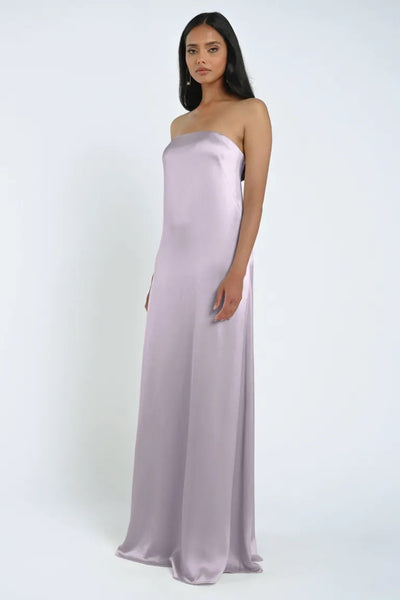 A woman stands wearing a James Bridesmaid Dress by Jenny Yoo from Bergamot Bridal, featuring a strapless neckline and flowing floor-length design in pastel lilac luxe satin fabric.