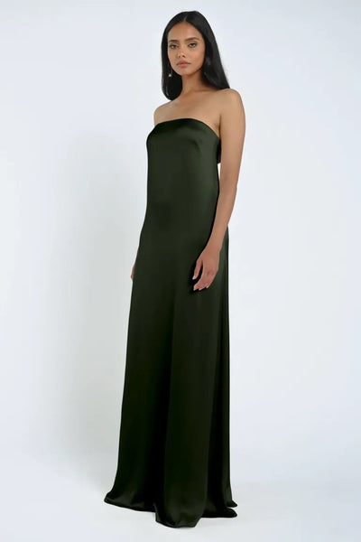 A woman stands wearing the James Bridesmaid Dress by Jenny Yoo from Bergamot Bridal, a strapless, floor-length dark green gown made of luxe satin fabric, against a plain white background.