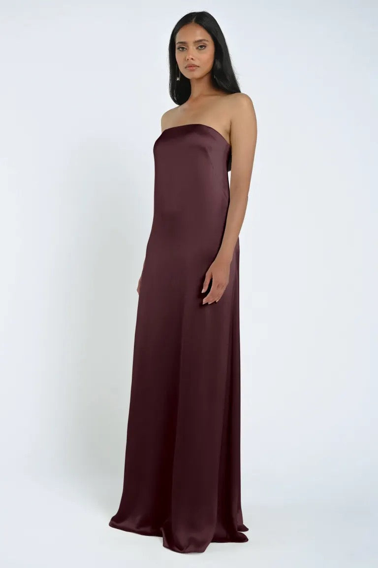 A woman stands against a plain background, wearing the James Bridesmaid Dress by Jenny Yoo from Bergamot Bridal, made of luxe satin fabric and featuring a strapless, floor-length maroon design.