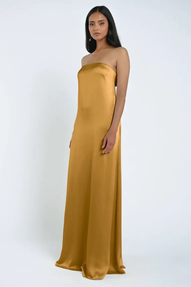 A person stands wearing the James Bridesmaid Dress by Jenny Yoo from Bergamot Bridal, a strapless, floor-length gown in mustard yellow crafted from luxe satin fabric against a plain white background.