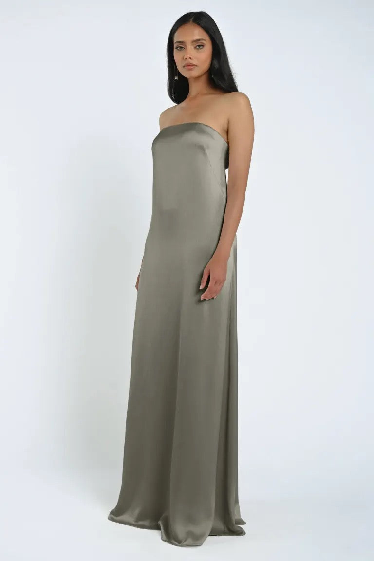 A woman stands wearing an elegant, floor-length James Bridesmaid Dress by Jenny Yoo in a neutral color, crafted from luxe satin fabric. Her hair is straight and she faces the camera with a neutral expression against a plain background. The dress is from the Bergamot Bridal collection.