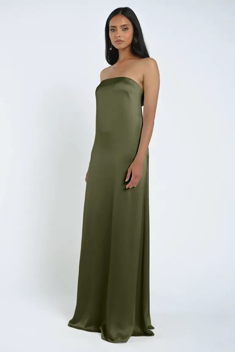 A person wearing an olive green James Bridesmaid Dress by Jenny Yoo from Bergamot Bridal, featuring a strapless neckline and floor-length luxe satin fabric, stands against a plain white background.