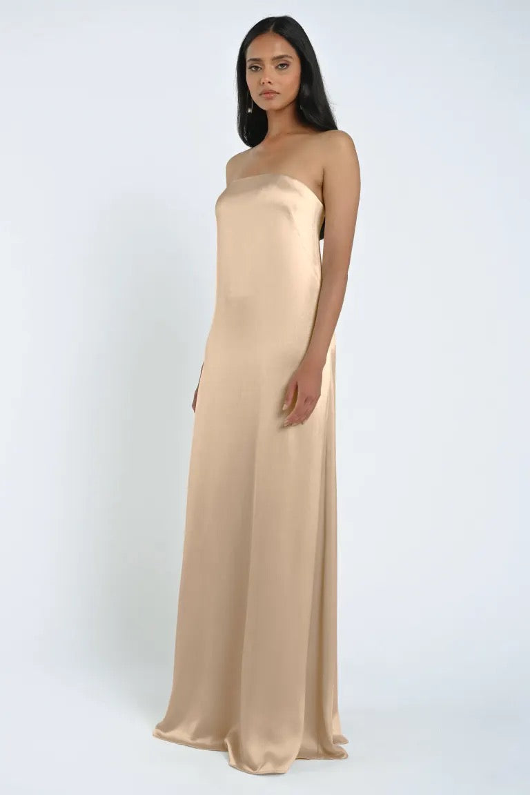 A woman stands wearing a long, strapless James Bridesmaid Dress by Jenny Yoo, made from luxe satin fabric with a sheath silhouette against a plain white background.