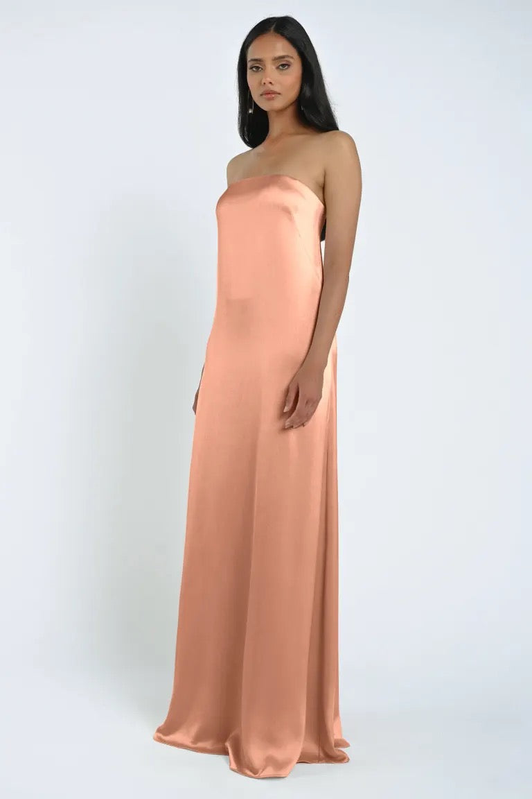 A person stands looking forward, wearing a strapless, floor-length James Bridesmaid Dress by Jenny Yoo with a sheath silhouette in luxe satin fabric from Bergamot Bridal against a plain white background.