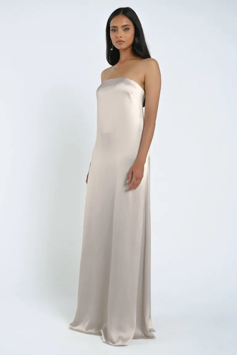 A woman is standing against a plain background, wearing the James Bridesmaid Dress by Jenny Yoo from Bergamot Bridal, a floor-length masterpiece with a strapless neckline crafted from luxe satin fabric.