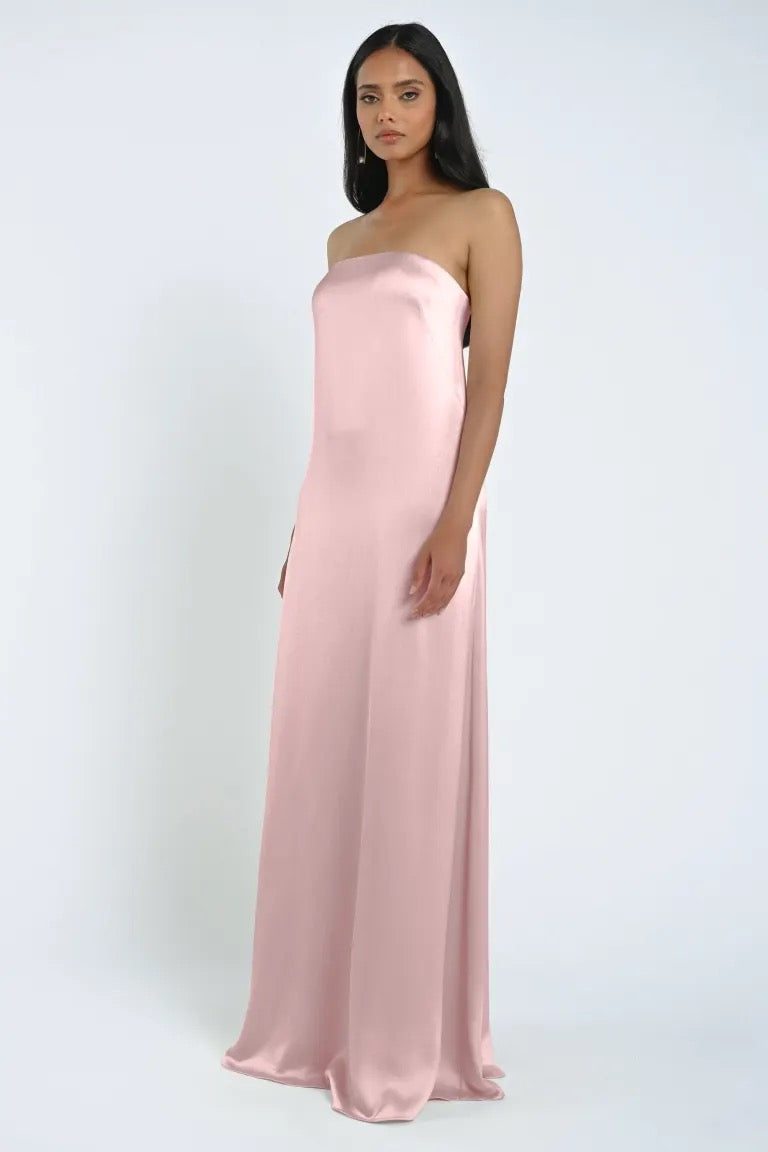 A woman stands wearing the James Bridesmaid Dress by Jenny Yoo for Bergamot Bridal, a long, strapless gown made from luxe satin fabric in a soft pink hue, against a plain background.