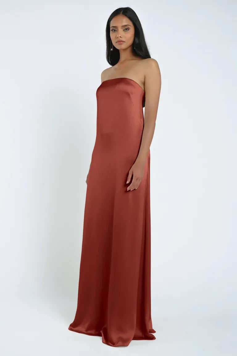 A woman stands wearing a strapless, floor-length James Bridesmaid Dress by Jenny Yoo made from luxe satin fabric in a stunning rust color against a plain white background.