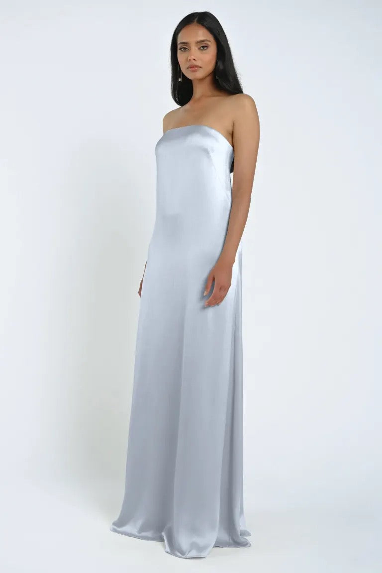 A woman stands against a plain background wearing a strapless James Bridesmaid Dress by Jenny Yoo, designed by Bergamot Bridal, made of floor-length, light blue luxe satin fabric.