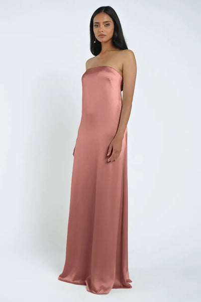 A woman wearing a luxe satin James Bridesmaid Dress by Jenny Yoo in rose pink from Bergamot Bridal stands against a plain white background.