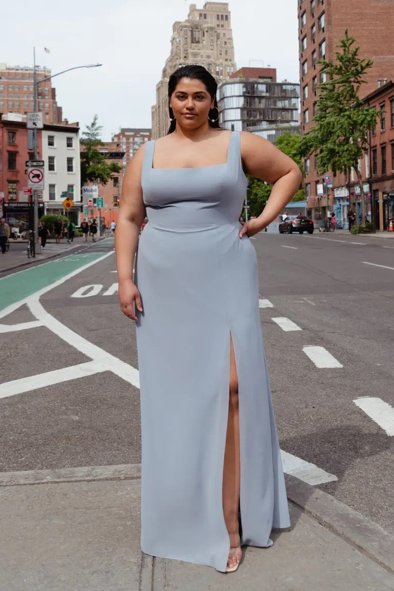 A person wearing a sleeveless grey Jenna - Bridesmaid Dress by Jenny Yoo chiffon bridesmaid dress with a thigh-high side slit and a square neckline standing on a city street.