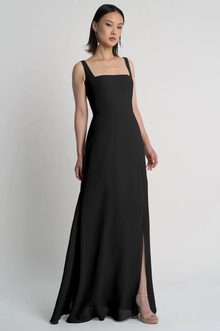 A woman in an elegant black sleeveless Jenna - Bridesmaid Dress by Jenny Yoo featuring a square neckline and a side slit stands against a neutral background from Bergamot Bridal.