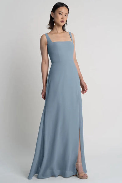 Woman in a blue Jenna chiffon bridesmaid dress by Jenny Yoo with a square neckline, standing against a plain background from Bergamot Bridal.