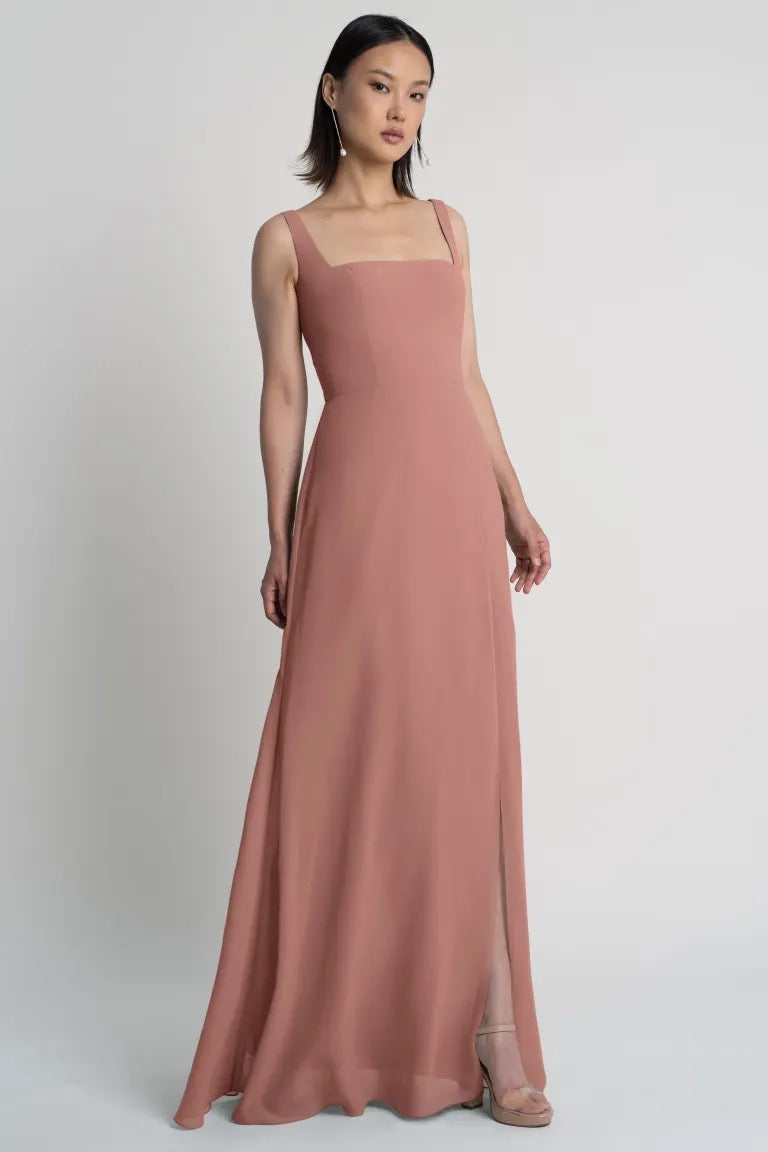 Woman posing in a sleeveless, floor-length, blush-colored Jenna Bridesmaid Dress by Jenny Yoo from Bergamot Bridal with a side slit.
