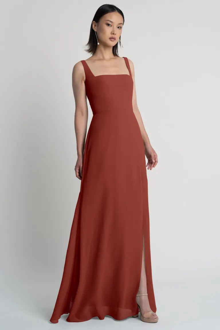 A woman modeling a long, rust-colored chiffon bridesmaid dress with square neckline and wide straps against a plain background by Bergamot Bridal.
