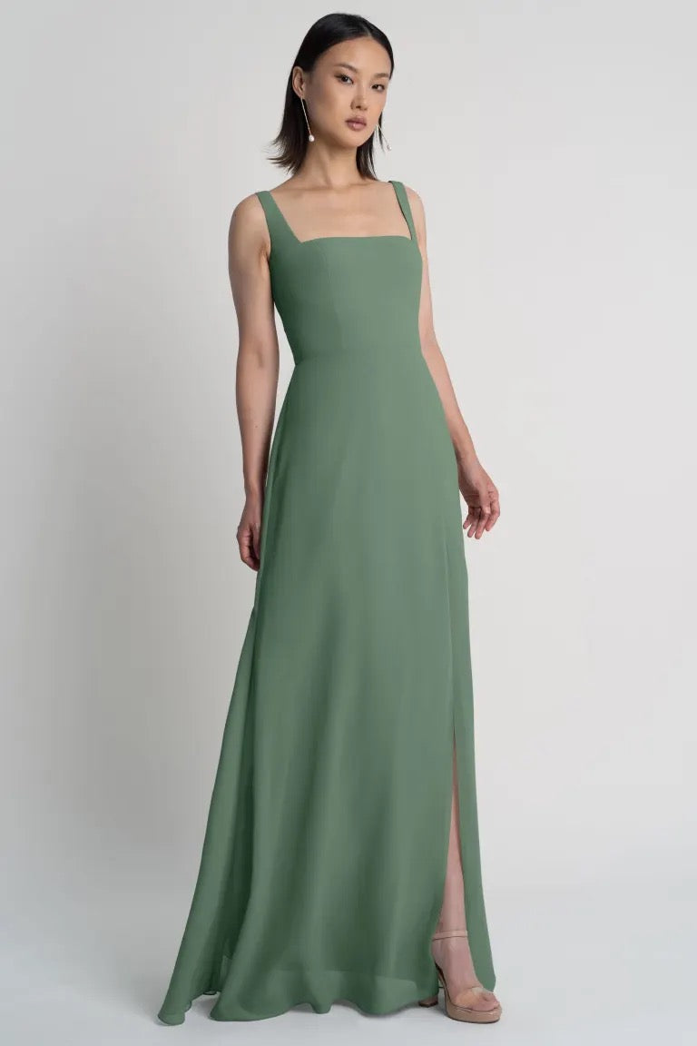 A woman wearing a sleeveless green Jenna chiffon bridesmaid dress with a square neckline stands against a neutral background. 
Product Name: Jenna - Bridesmaid Dress by Jenny Yoo
Brand Name: Bergamot Bridal