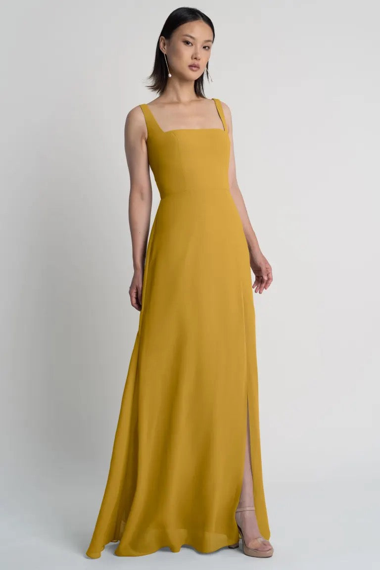 Woman in a Jenna bridesmaid dress by Jenny Yoo in mustard yellow with a square neckline standing against a neutral background, available at Bergamot Bridal.