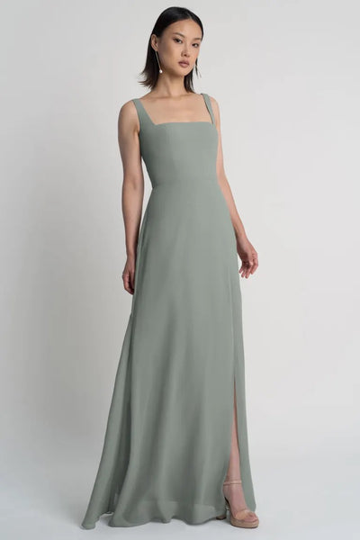 A woman in a long, sage green Jenna chiffon bridesmaid dress with a sleek silhouette and square neckline by Jenny Yoo, standing against a neutral background.