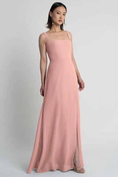 A woman in a Jenna - Bridesmaid Dress by Jenny Yoo in pale pink with a square neckline standing against a grey background from Bergamot Bridal.