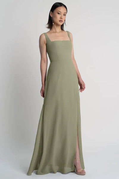 A woman in an elegant olive green sleeveless Jenna chiffon bridesmaid dress by Jenny Yoo with a side slit stands against a plain background.