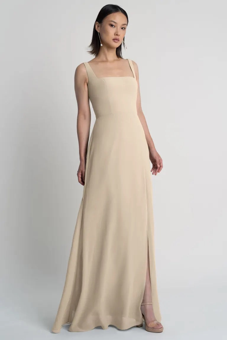 A woman poses in a minimalist beige Jenna chiffon bridesmaid dress by Jenny Yoo with a square neckline against a plain background from Bergamot Bridal.