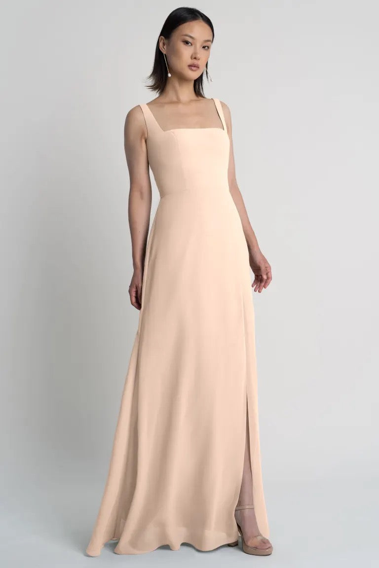 A woman modeling a long, pale beige sleeveless Jenna bridesmaid dress by Jenny Yoo with a simple, elegant design and square neckline from Bergamot Bridal.