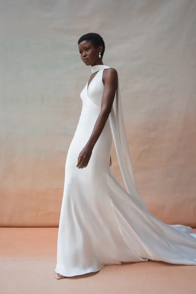 A person poses in the Jennifer Wedding Dress by Jenny Yoo from Bergamot Bridal, showcasing a floor-length, fitted white bridal gown with a train and shoulder drape in luxe satin fabric. The background transitions from light brown to beige.