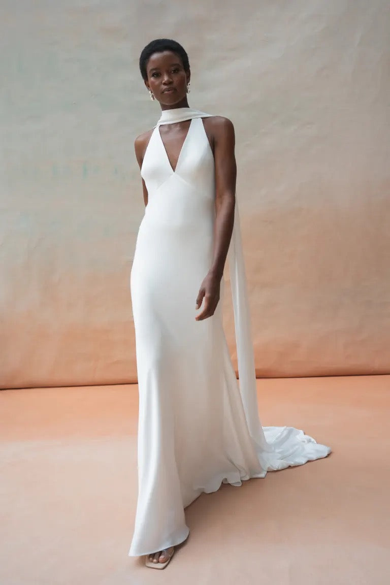 A person wearing the Jennifer Wedding Dress by Jenny Yoo from Bergamot Bridal, featuring a sleeveless luxesatin design with a plunging V-neckline and a long scarf, walks against a simple backdrop.