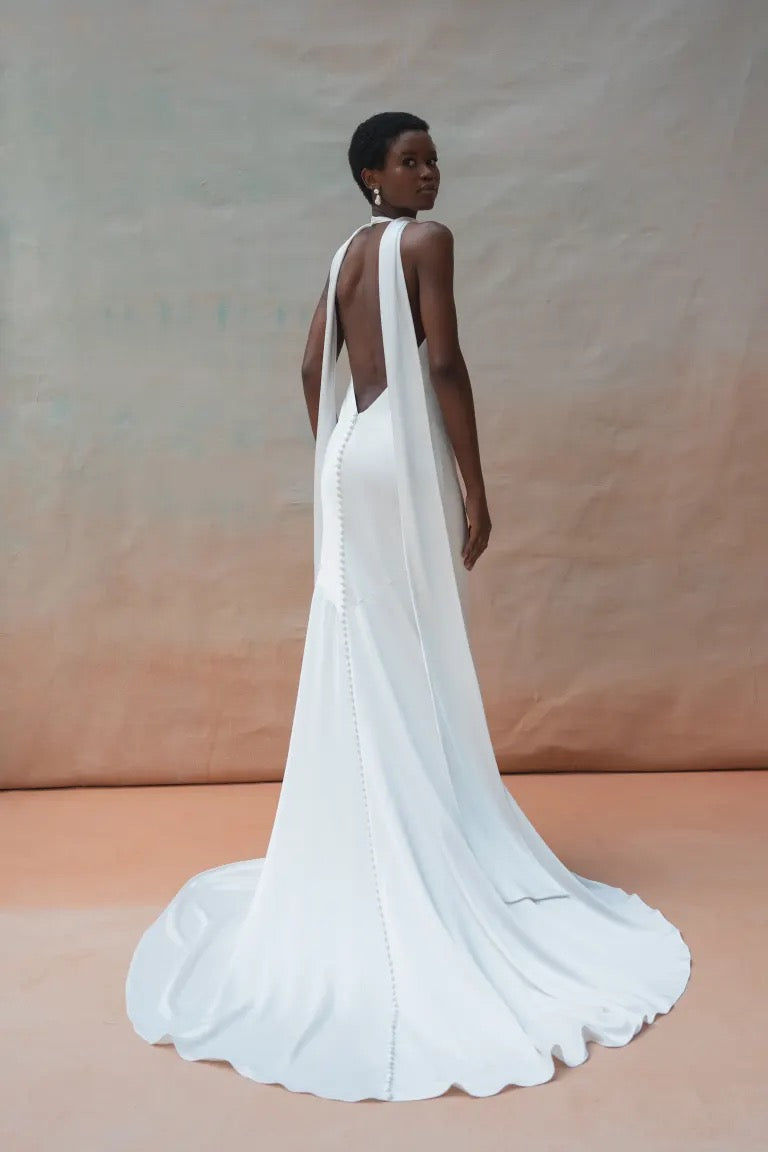 A person wearing the Jennifer Wedding Dress by Jenny Yoo, a floor-length, white satin bridal gown with a plunging V-neck from Bergamot Bridal, poses against a neutral backdrop, looking over their shoulder.