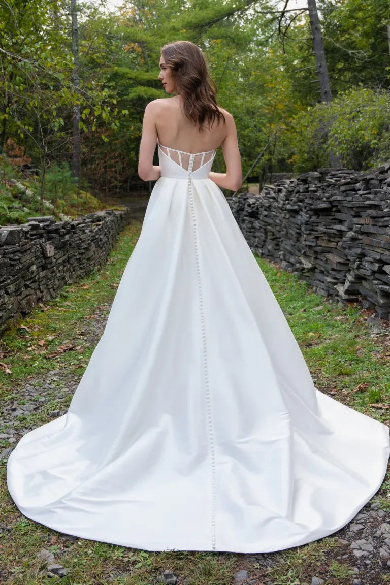 A woman in a white bridal gown with an open back design and an A-line skirt standing in a wooded area wearing the Mollie wedding dress by Jenny Yoo from Bergamot Bridal.