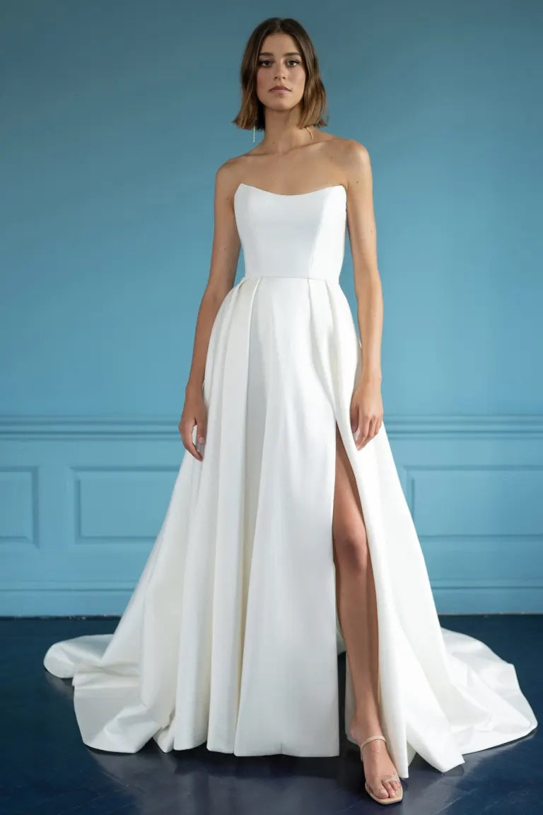 A woman in an elegant white strapless Mollie - Jenny Yoo Wedding Dress with a high slit standing against a blue backdrop from Bergamot Bridal.