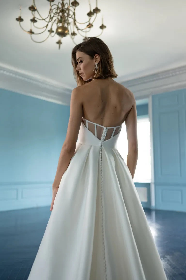A woman in an elegant white bridal gown with an A-line skirt and an open back stands in a room with light blue walls and a chandelier wearing the Mollie wedding dress from Bergamot Bridal.