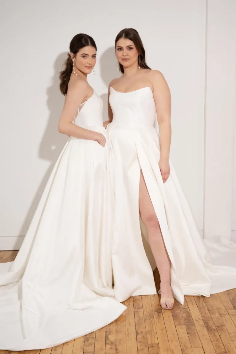 Two women in elegant white Mollie - Jenny Yoo wedding dresses from Bergamot Bridal with scoop necklines and a thigh-high slit posing together.