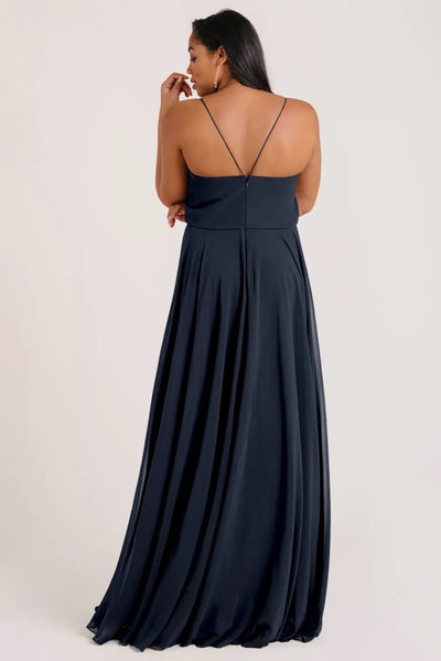A woman in an elegant navy blue Inesse bridesmaid dress by Jenny Yoo with a V-neck and a crossed strap design at the back, standing against a neutral background from Bergamot Bridal.