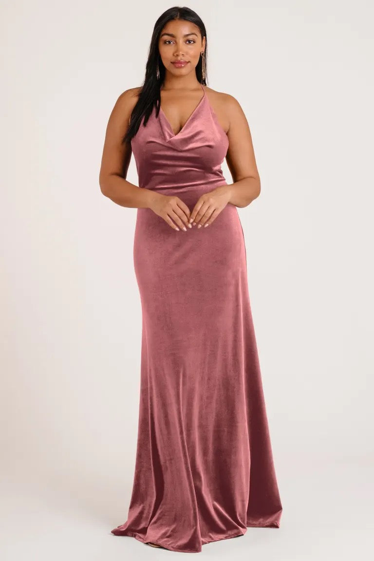 A woman poses in an elegant rose-colored velvet Sullivan bridesmaid dress by Jenny Yoo with a halter cowl neckline from Bergamot Bridal.
