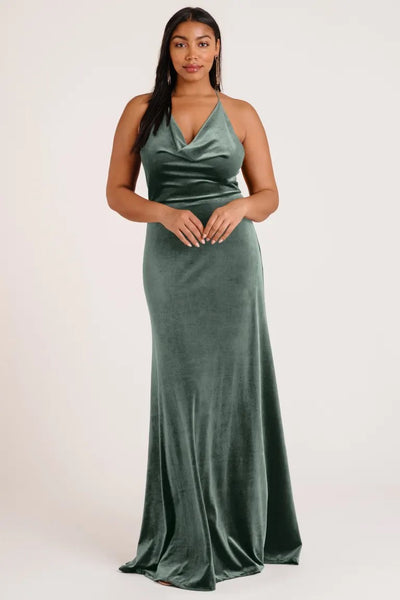 A woman in an elegant green velvet Sullivan bridesmaid dress by Jenny Yoo with a halter cowl neckline standing against a light background from Bergamot Bridal.