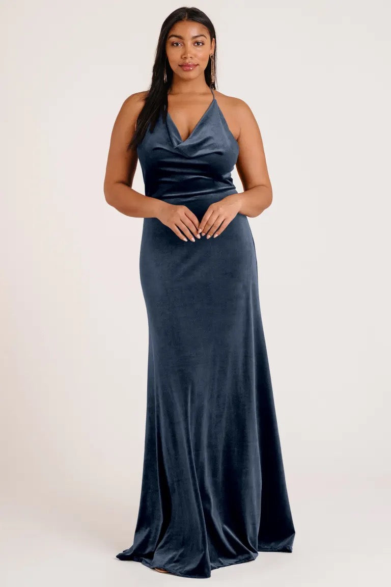 Woman in an elegant blue velvet Sullivan - Bridesmaid Dress by Jenny Yoo with a halter cowl neckline, posing against a neutral background from Bergamot Bridal.