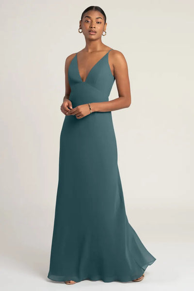 Person wearing the Jude - Bridesmaid Dress by Jenny Yoo, a sleeveless, teal-colored, floor-length dress with a deep V neckline and an empire waist seam, standing against a plain background.