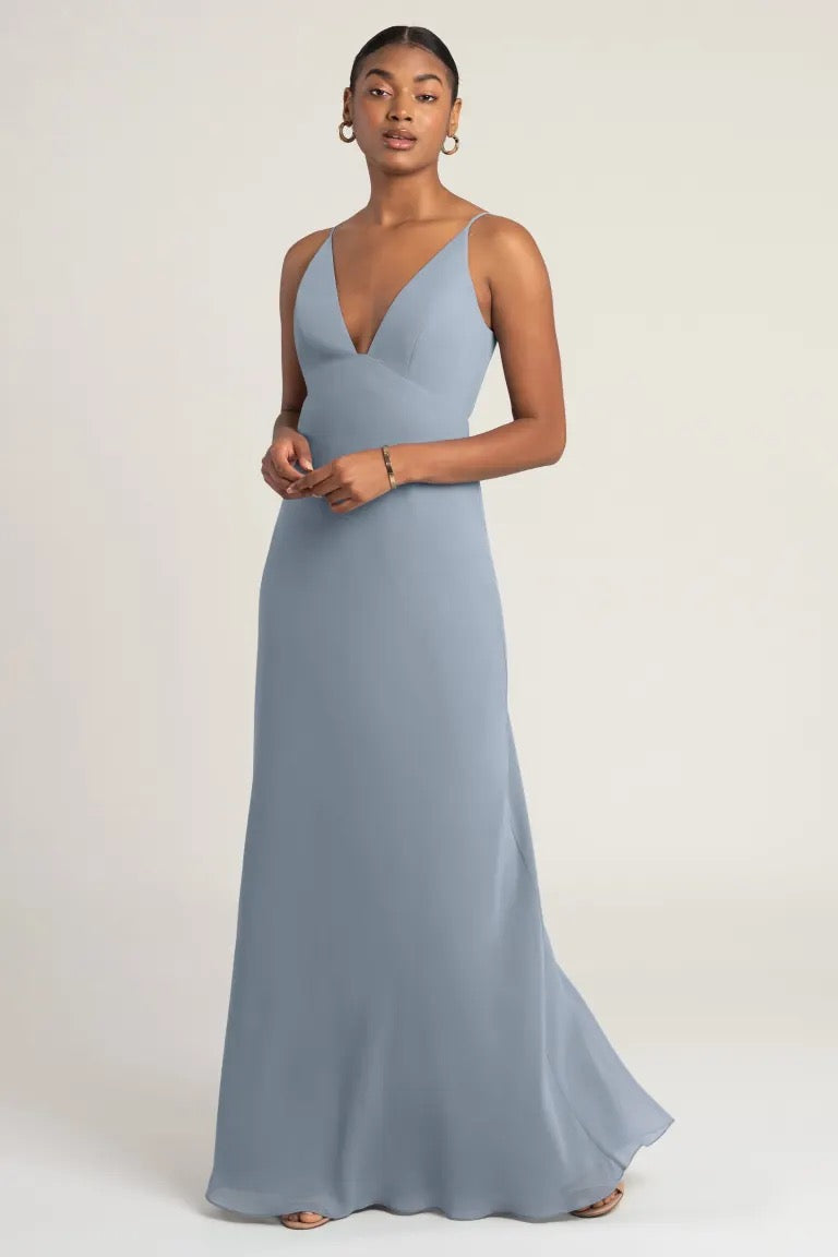 A woman in an elegant blue evening gown with a notched neckline posing against a neutral background, wearing the Jude Bridesmaid Dress by Jenny Yoo from Bergamot Bridal.