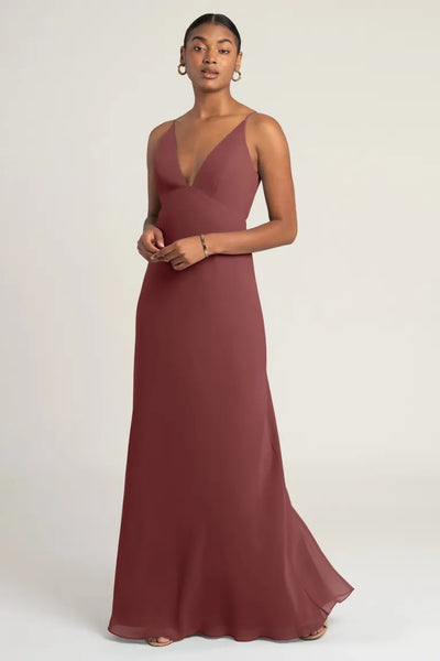 A woman in a Jude bridesmaid dress by Jenny Yoo from Bergamot Bridal, an elegant maroon chiffon evening gown with an empire waist, a v-neckline, and sleeveless design standing against a neutral background.