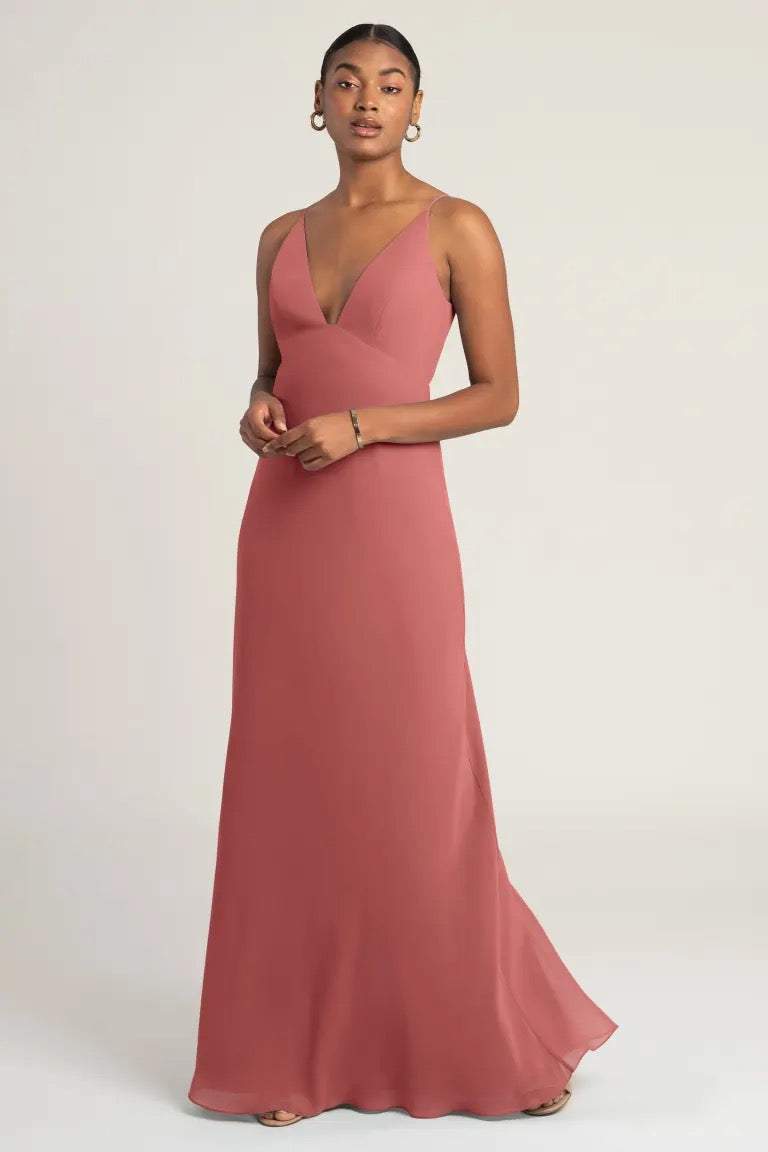 A woman in a Jude - Bridesmaid Dress by Jenny Yoo in an elegant rose-colored chiffon evening gown with an empire waist posing against a neutral background from Bergamot Bridal.