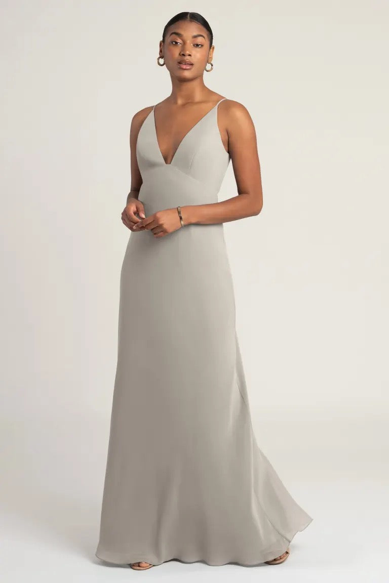 A woman in an elegant chiffon taupe Jude bridesmaid dress by Jenny Yoo posing against a neutral background from Bergamot Bridal.