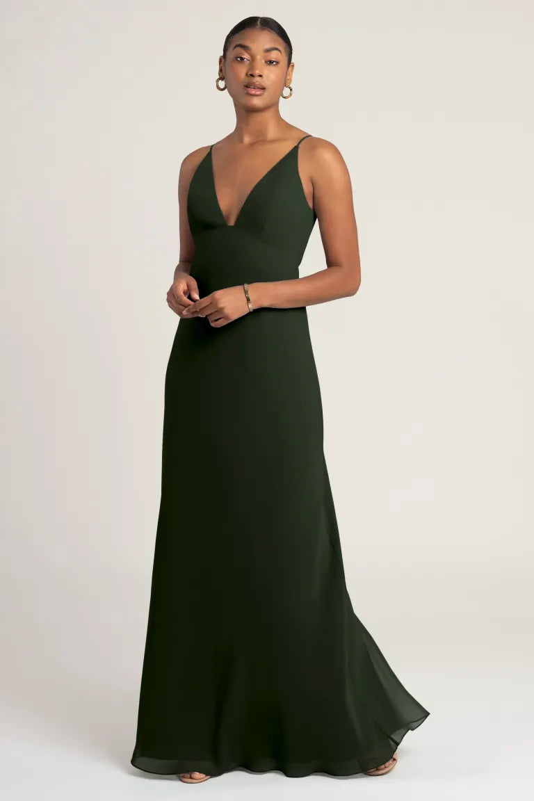 A woman wearing the Jude - Bridesmaid Dress by Jenny Yoo from Bergamot Bridal, a dark green, sleeveless, floor-length gown featuring a deep V-neckline and an empire waist seam, stands against a plain background. The body-skimming bias-cut skirt enhances her silhouette as she accessorizes with small hoop earrings and a bracelet.