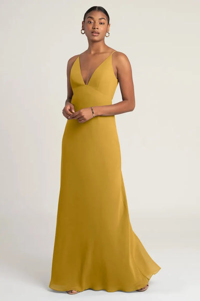 A woman in a Jude bridesmaid dress by Jenny Yoo in an elegant mustard yellow empire waist evening gown poses against a neutral backdrop.