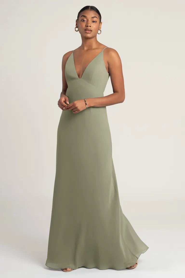 A woman in an elegant olive green empire waist Jude bridesmaid dress by Jenny Yoo with a notched neckline posing for the camera from Bergamot Bridal.